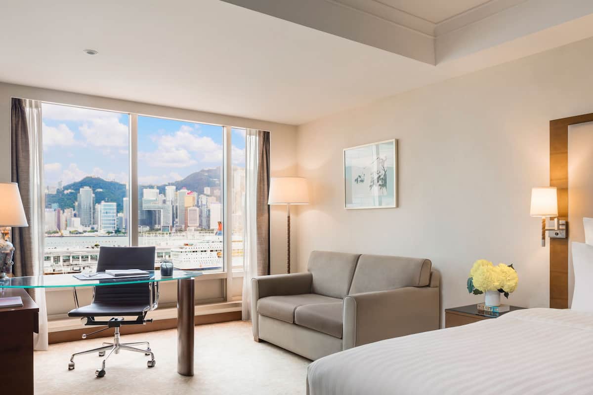 A hotel room with a large window, a bed, a sofa, a desk with a chair, and a nightstand with a lamp and flowers.