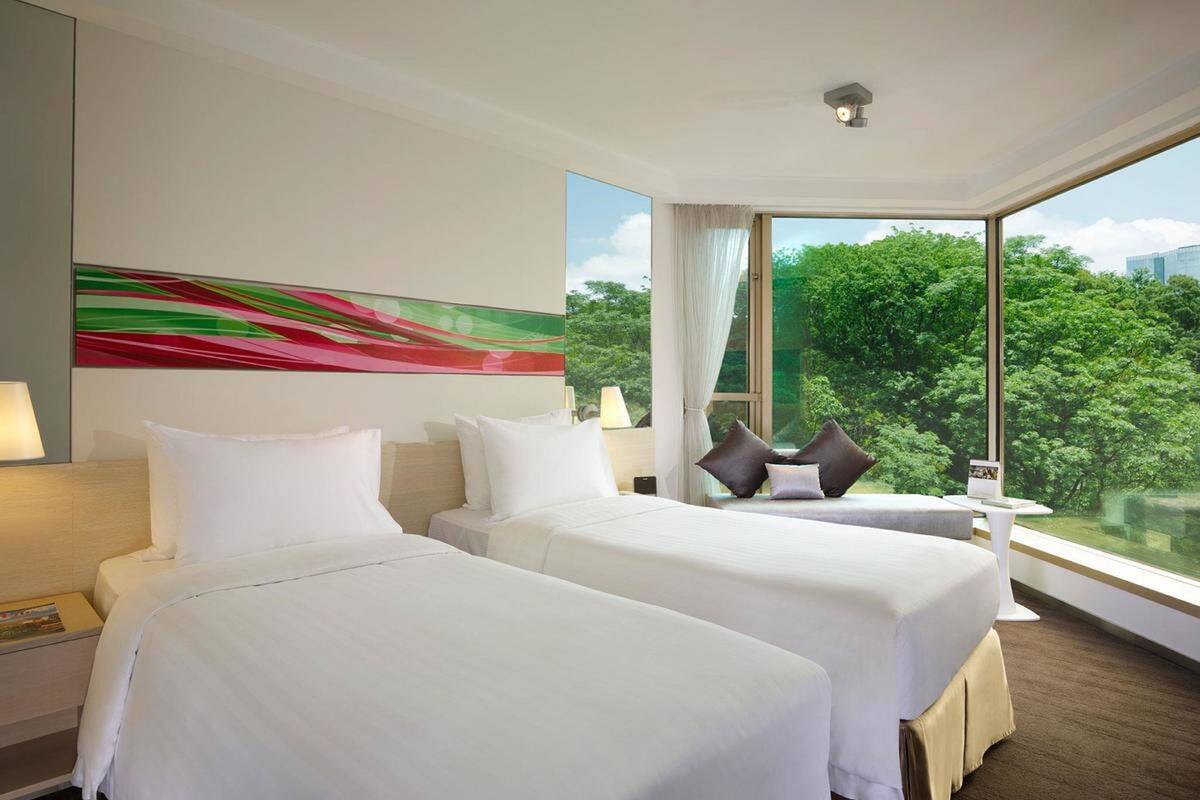 A hotel room with two beds, a large window with a view of greenery, and a colorful abstract painting on the wall.