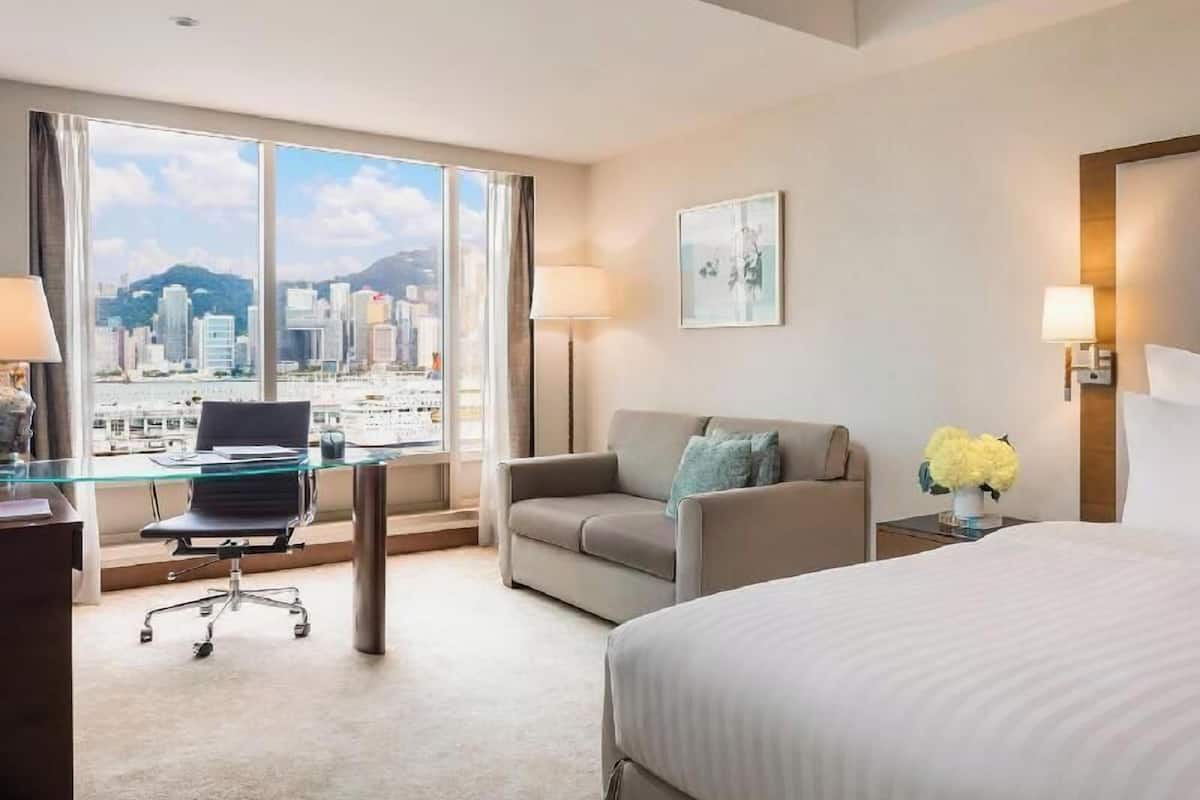 A hotel room with a large window offering a city view, a bed, a desk with a chair, and a sofa.