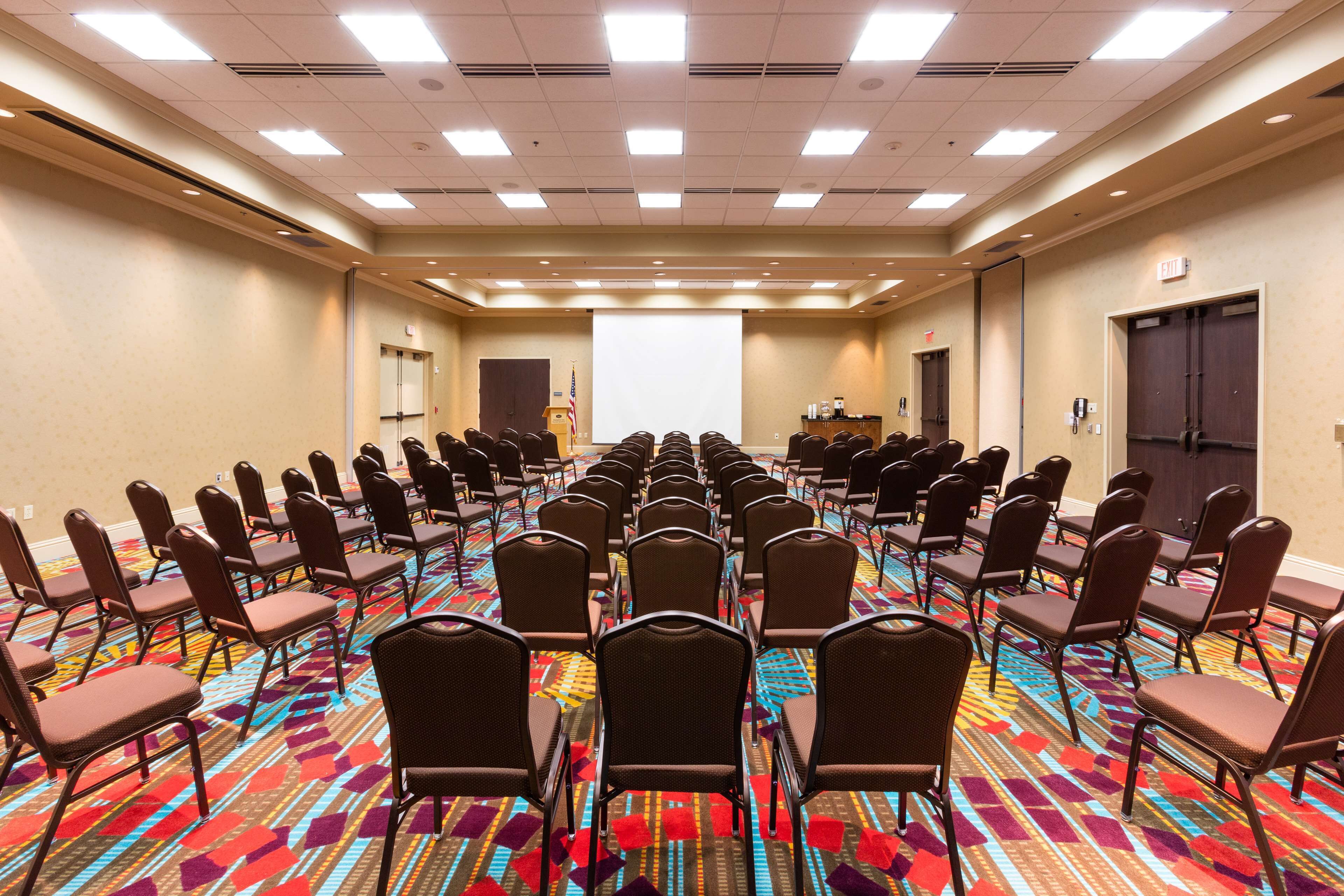 A conference room with rows of chairs facing a projection screen, carpeted floor, and a door with an exit sign.