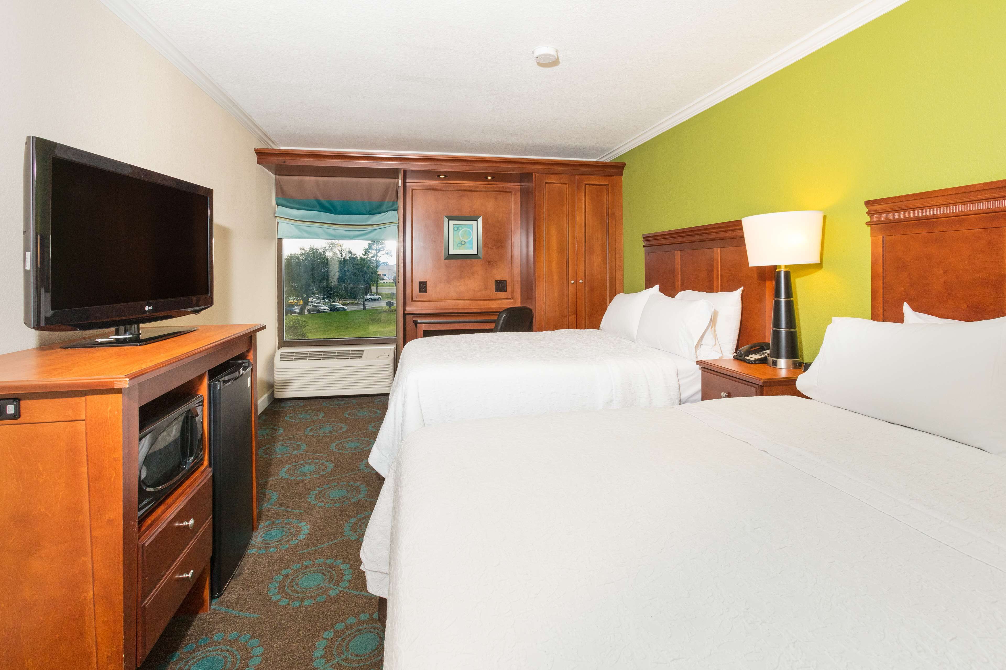 A hotel room with two beds, a television, a desk with a computer, and a window with a view of a green lawn.