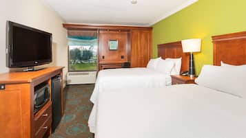 A hotel room with two beds, a television, a desk with a computer, and a window with a view of a green lawn.
