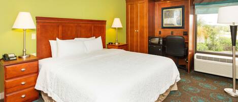 A hotel room with a large bed, wooden headboard, two bedside tables with lamps, a desk with a chair, and a window with a view of greenery.