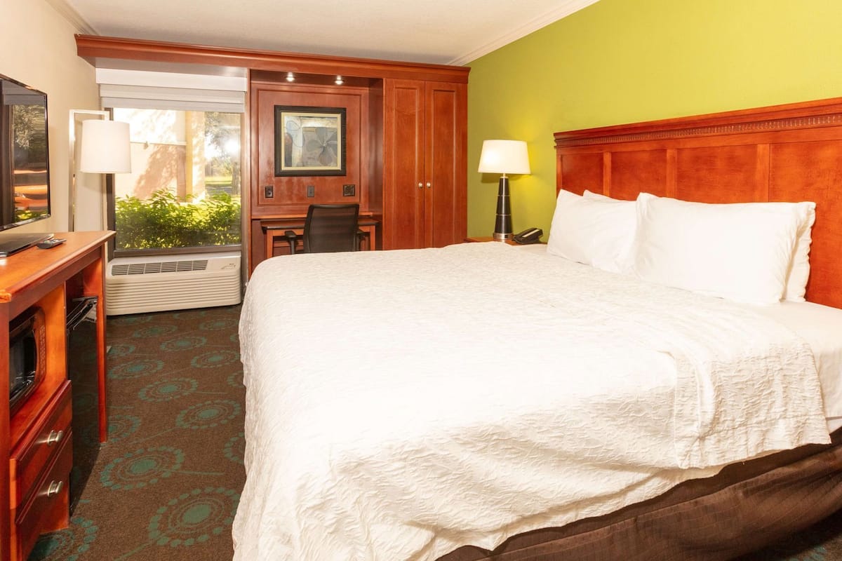 A hotel room with a large bed, a desk, a chair, a lamp, and a TV.