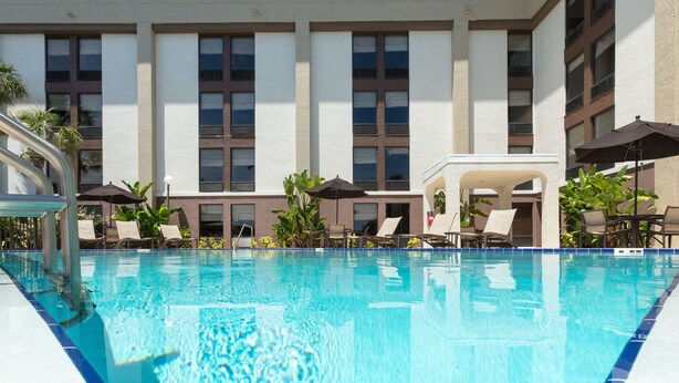 A swimming pool with lounge chairs and umbrellas, surrounded by a building with multiple windows and a tiled floor.