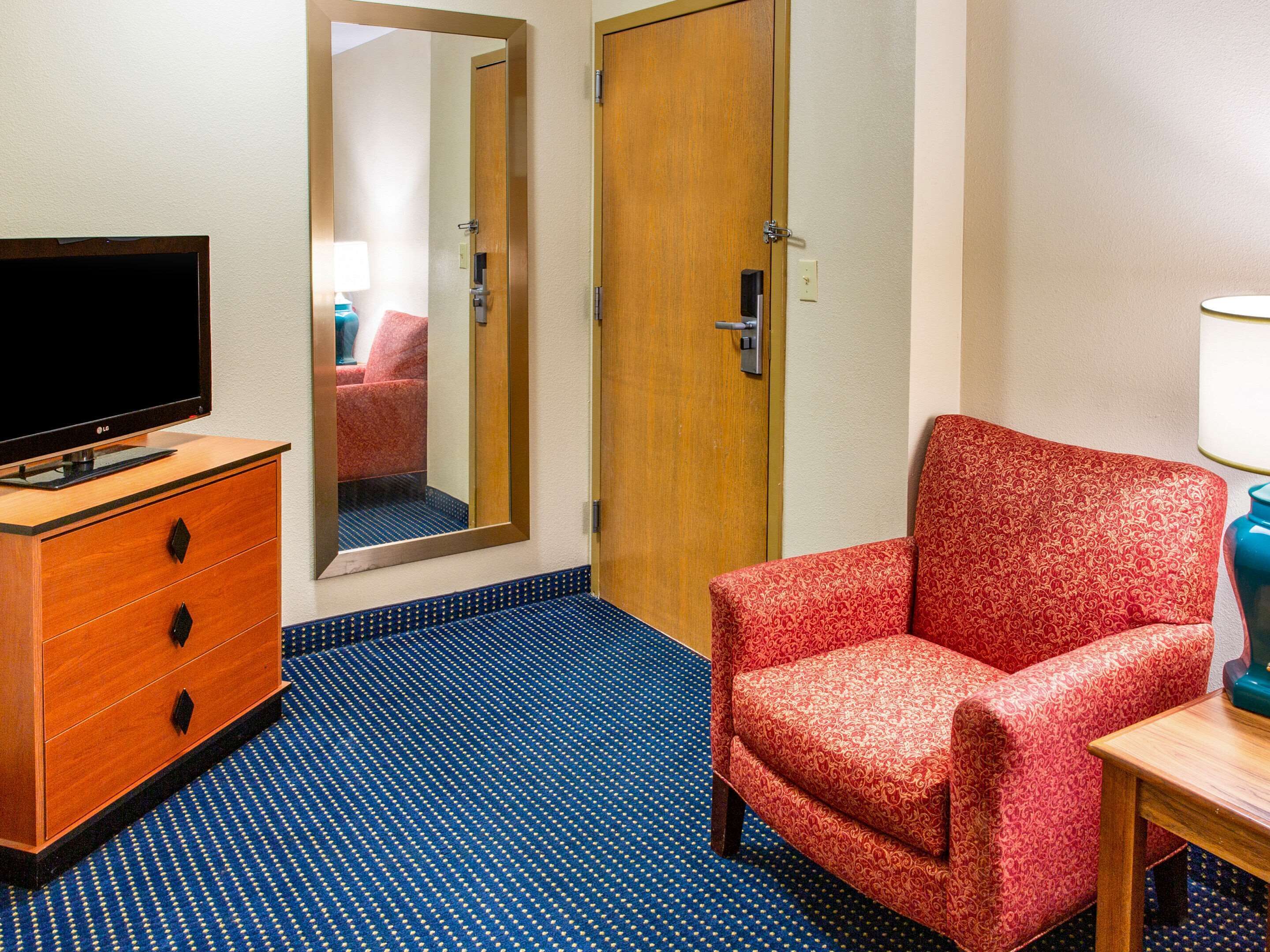 A hotel room with a wooden dresser, a television, a red patterned armchair, a mirror, and a blue carpet.