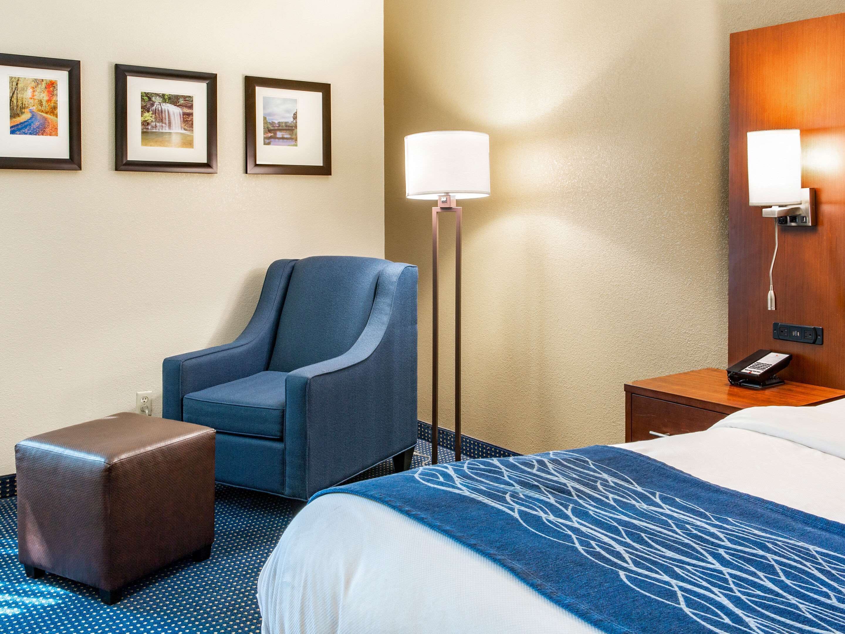 Quarto de hotel com cama, uma poltrona azul, um pequeno banquinho, mesa de cabeceira com luminária e três quadros na parede.