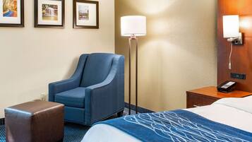 Quarto de hotel com cama, uma poltrona azul, um pequeno banquinho, mesa de cabeceira com luminária e três quadros na parede.