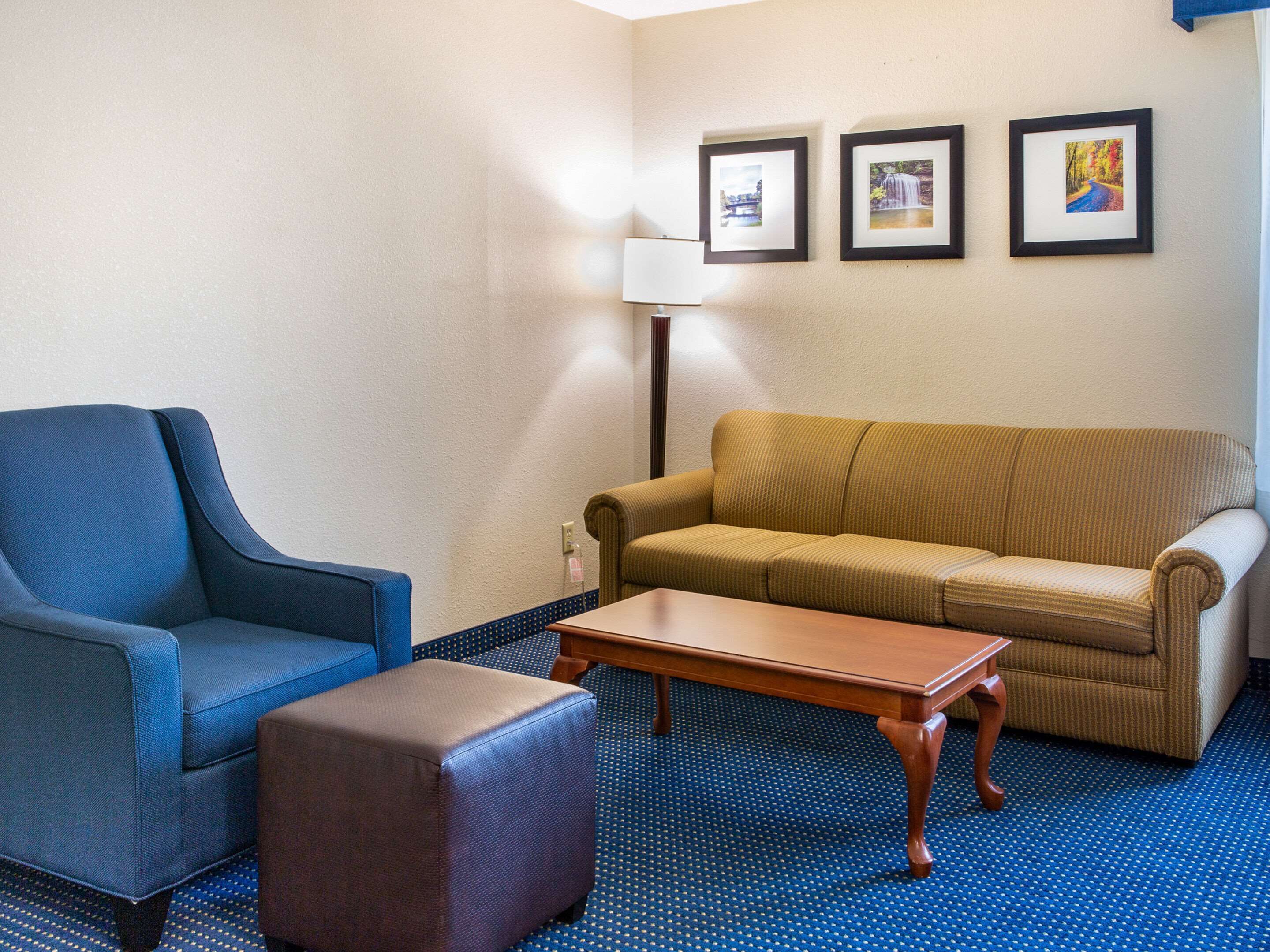 A hotel room with a blue armchair, a brown sofa, a wooden coffee table, and a blue patterned carpet. There are three framed pictures on the wall.