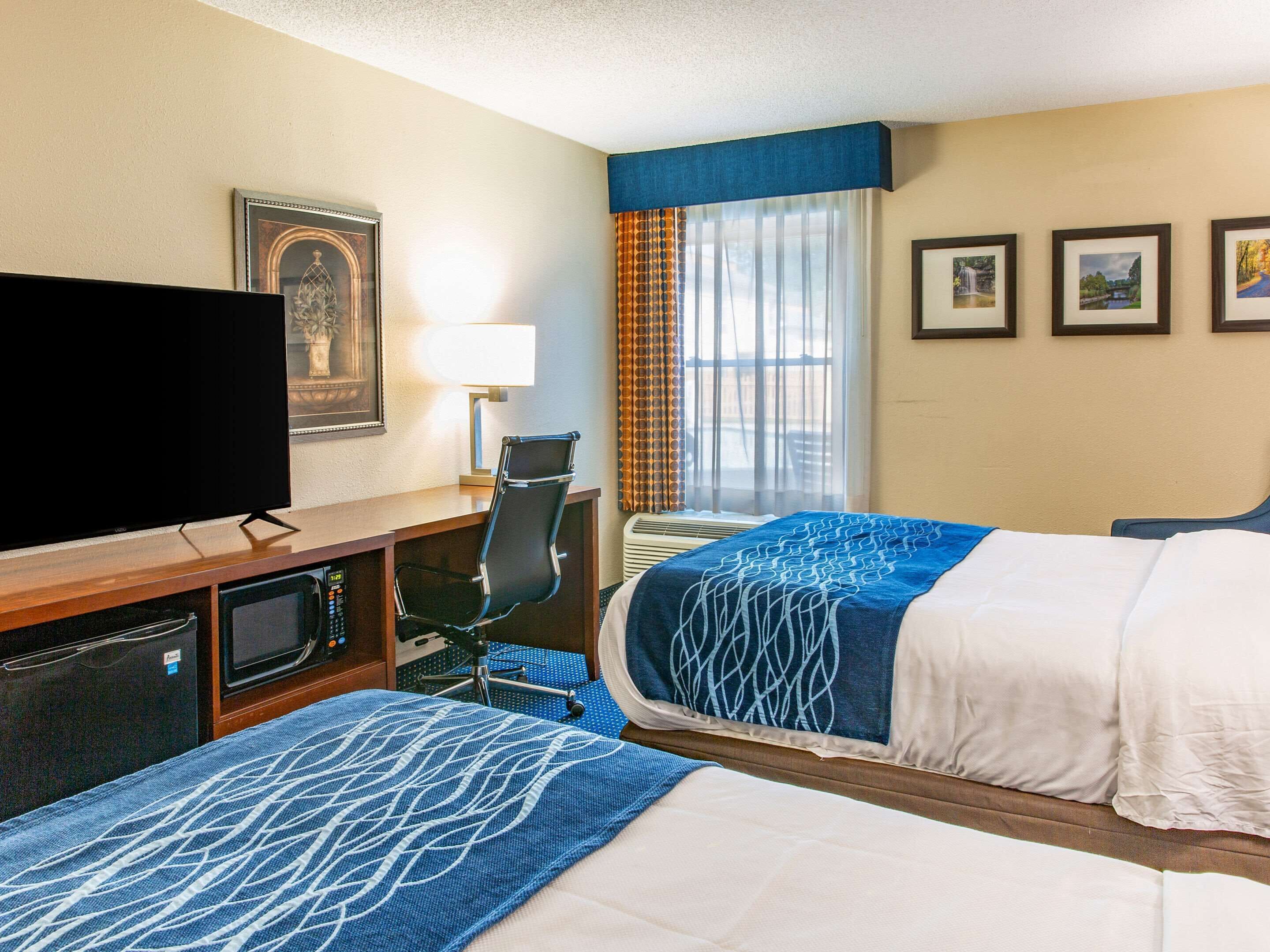 A hotel room with two beds, a desk with a TV, a chair, a desk lamp, and a window with curtains.