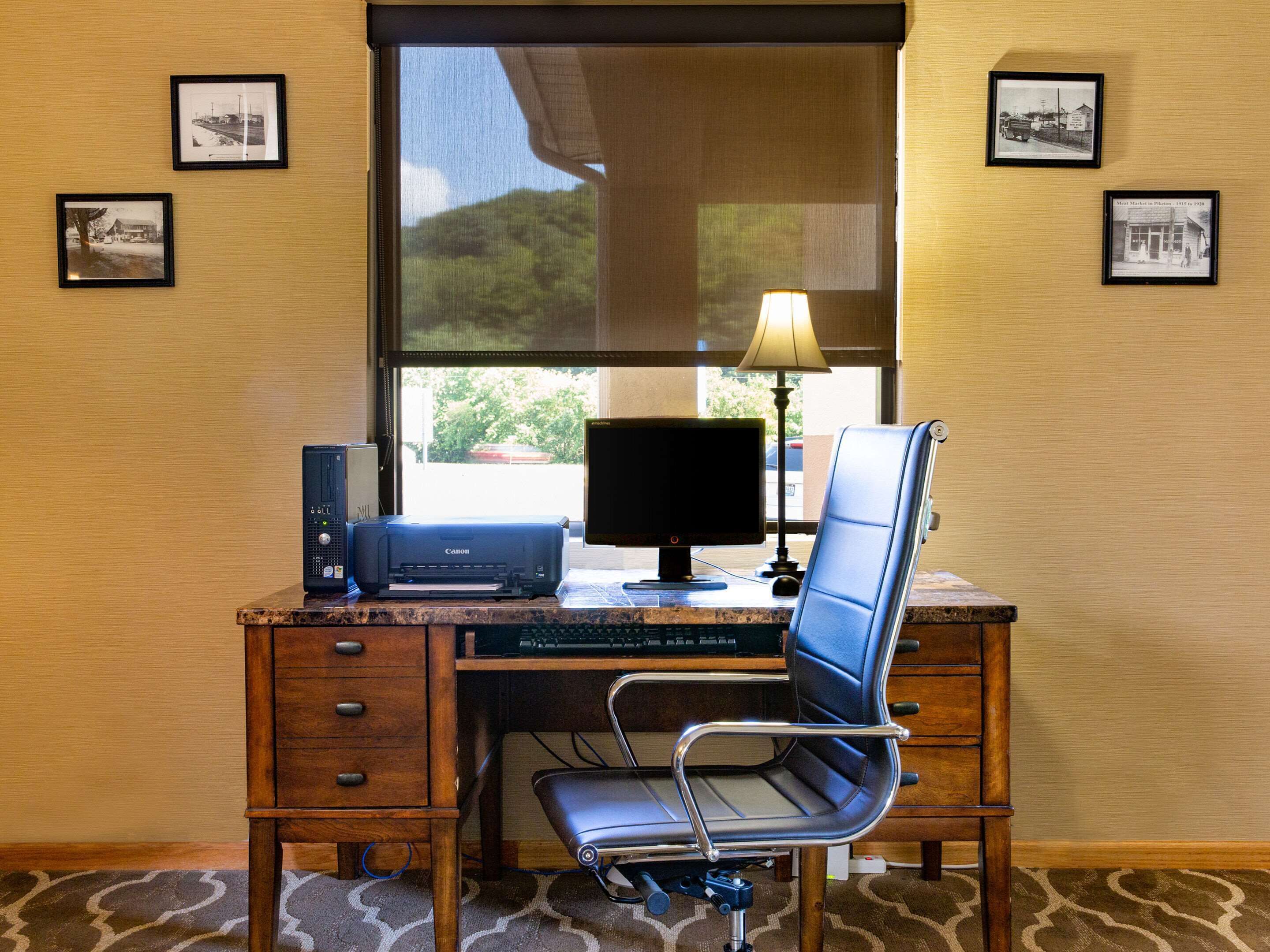 A wooden desk with a computer, monitor, and printer. There are framed pictures on the wall and a lamp on the desk.
