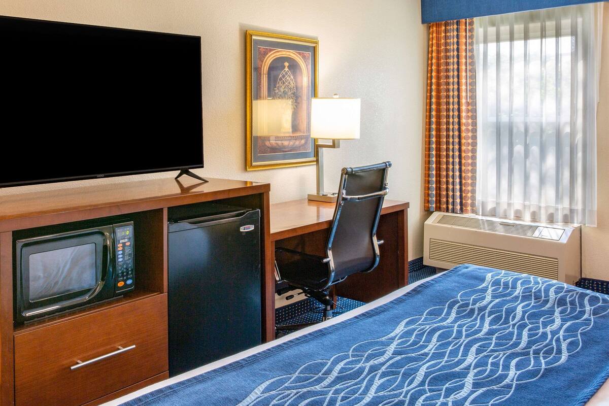 A hotel room with a bed, a desk with a computer, a television, a microwave, a mini-fridge, and a window with curtains.