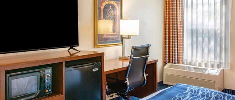 A hotel room with a bed, a desk with a computer, a television, a microwave, a mini-fridge, and a window with curtains.