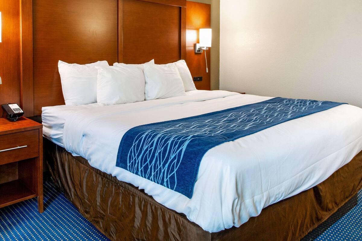 A hotel room with a large bed, a wooden headboard, a bedside table with a phone, and a wall-mounted lamp.