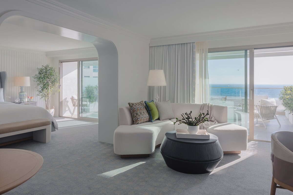 A modern hotel room with a large bed, a sofa, a coffee table, and a balcony with a view of the sea.
