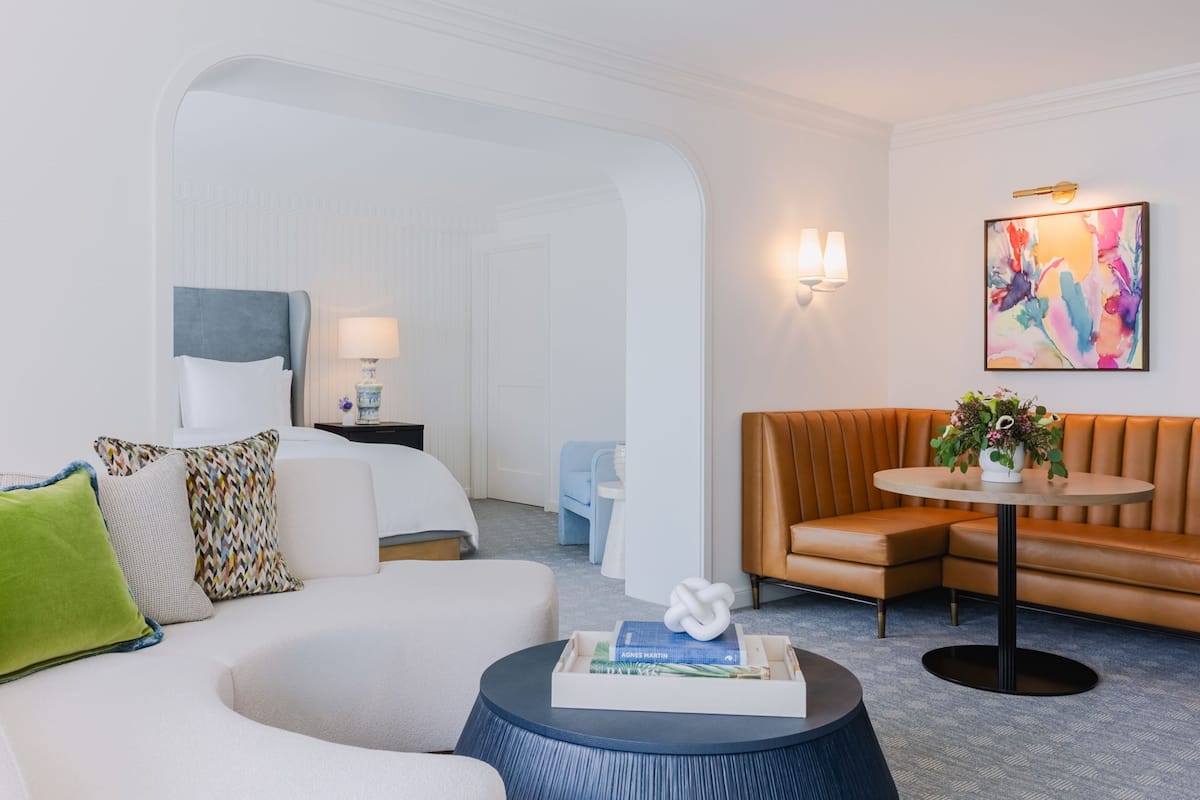 A modern hotel room with a white sofa, a round coffee table with books, a brown leather couch, a small round table with a plant, and a colorful abstract painting on the wall.