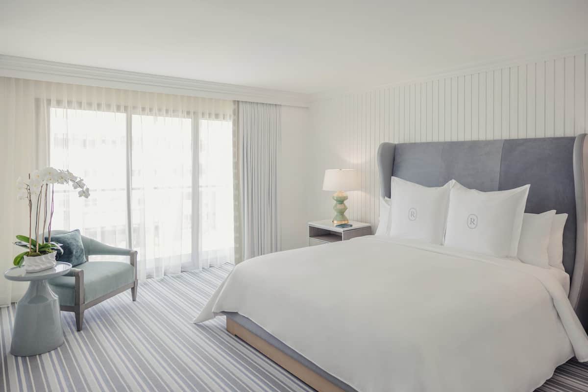 A hotel room with a large bed, a bedside table with a lamp, a chair, and a potted plant.