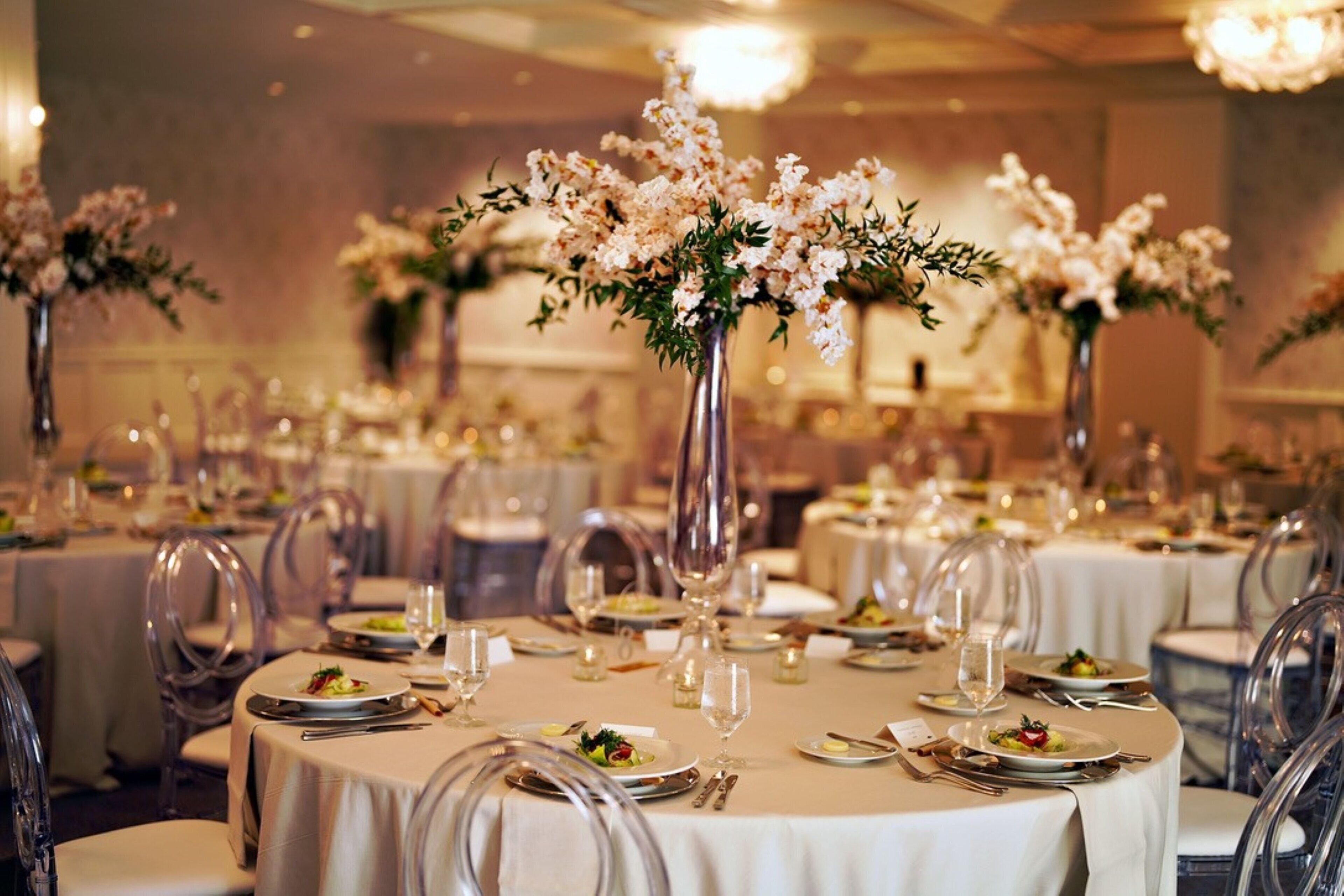 Un salón de banquetes con mesas redondas, sillas transparentes y centros de mesa florales.