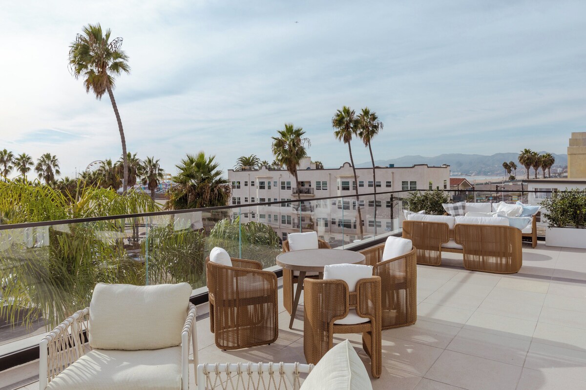 A rooftop terrace with wicker furniture, palm trees, and a view of a cityscape.
