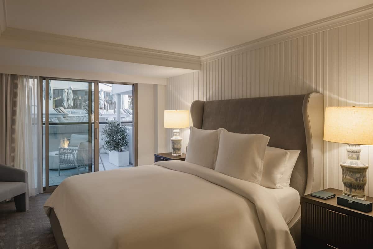 A hotel room with a large bed, two bedside tables with lamps, a sofa, and a sliding glass door leading to an outdoor area with a fire pit.