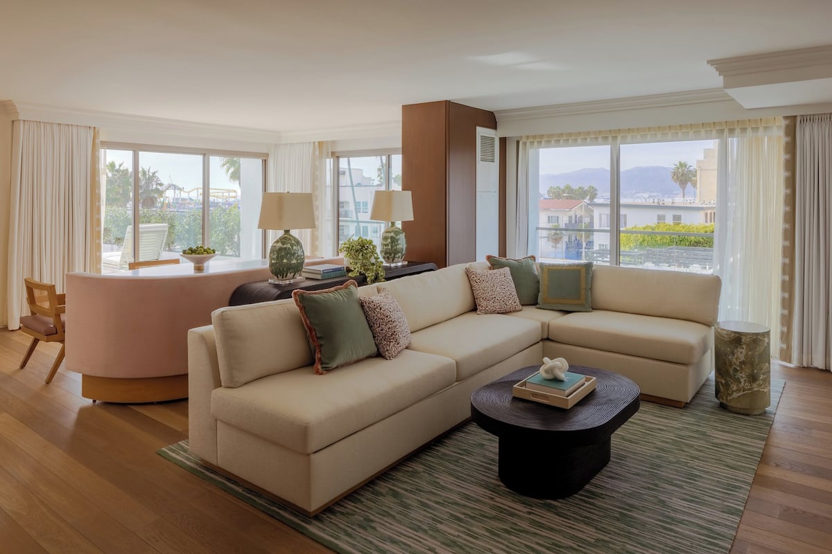 A modern hotel room with a large beige sectional sofa, a small round coffee table, and a dining area with a pink table and chairs.