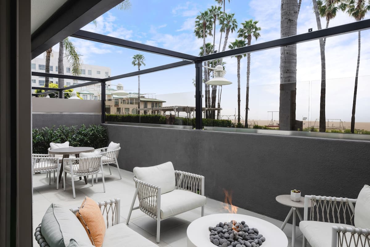 A modern outdoor seating area with white wicker furniture, a fire pit, and a view of palm trees and buildings.
