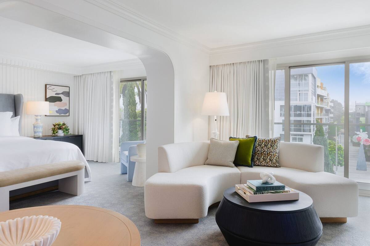 A modern hotel room with a large bed, a sofa, a round coffee table, and a view of the city through large windows.