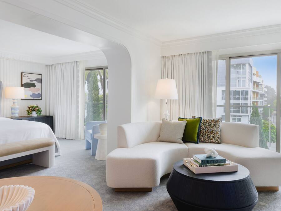 A modern hotel room with a large bed, a sofa, a round coffee table, and a view of the city through large windows.