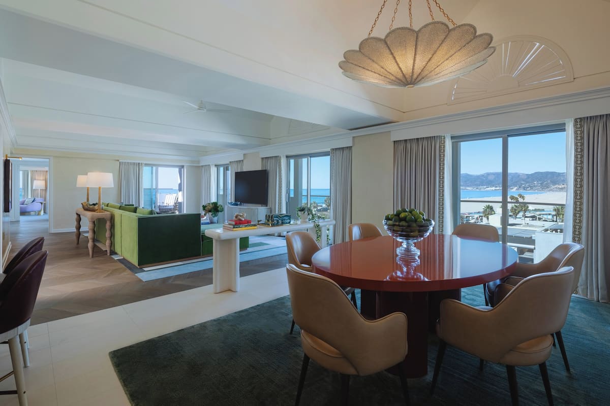 A spacious hotel room with a dining area, a green sofa, a TV, and a view of the beach.