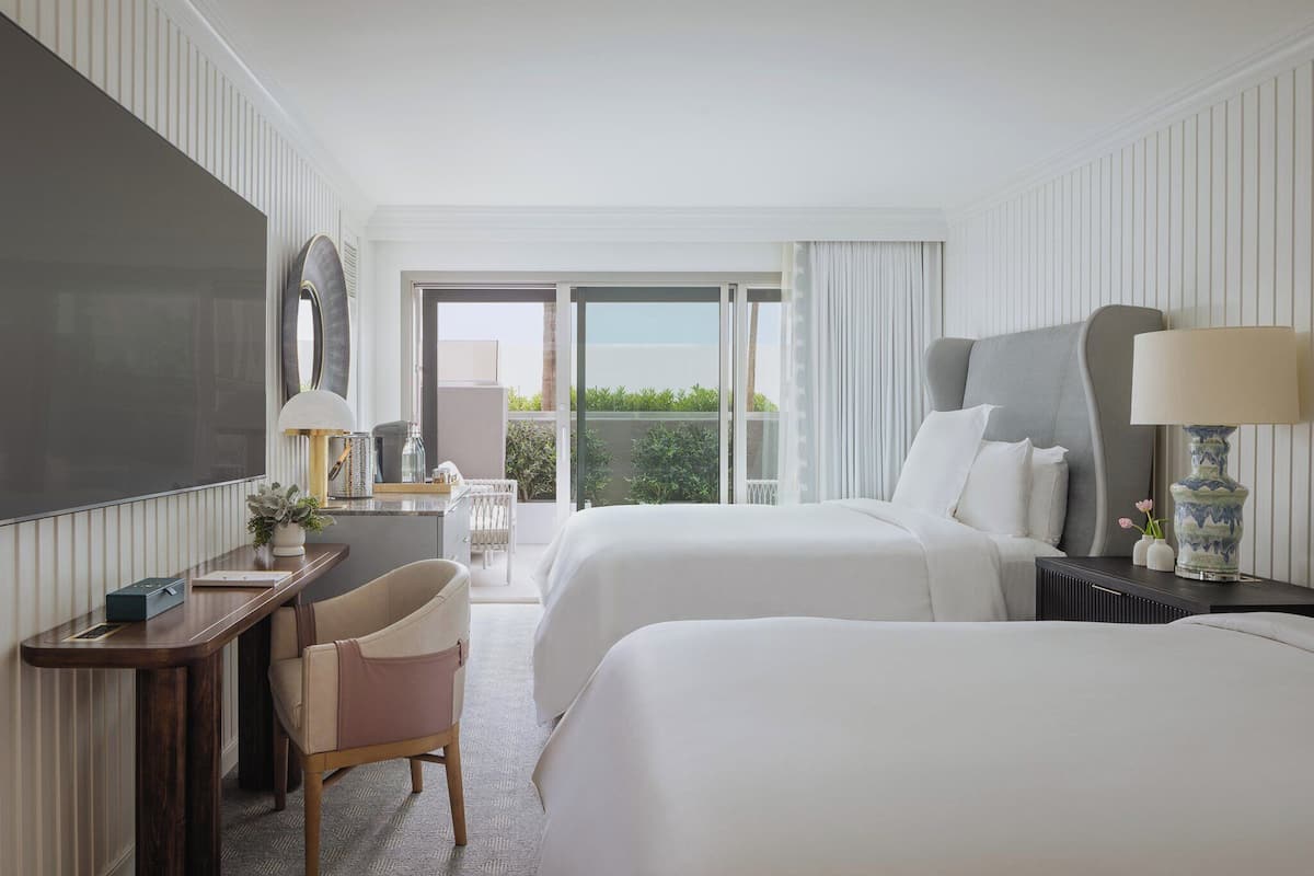 A hotel room with two beds, a desk, a chair, a TV, and a view of greenery outside.