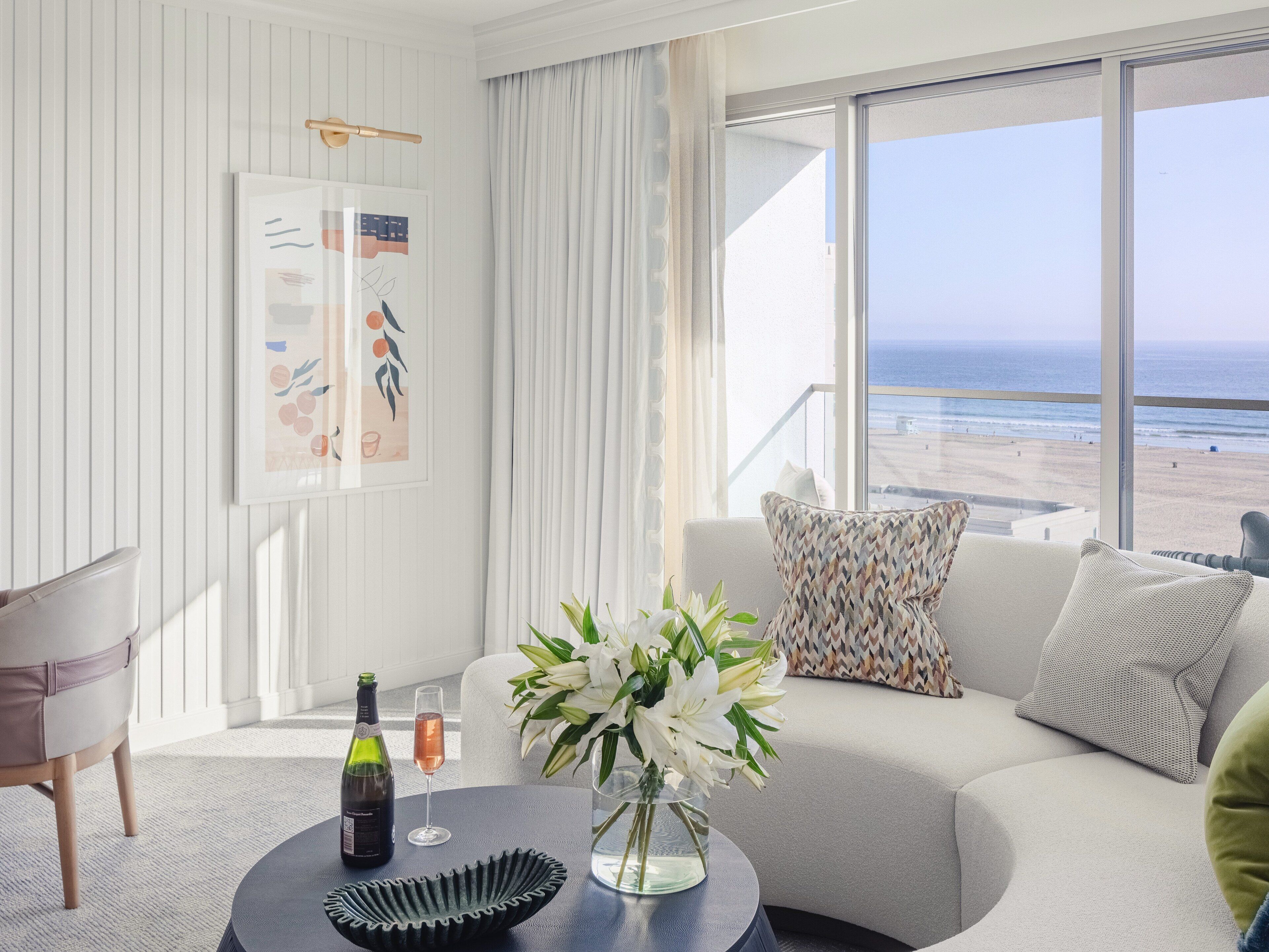 A modern living room with a view of the beach, a framed artwork, a bottle of wine, and a vase of flowers on a round table.