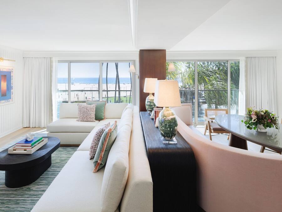 A modern hotel room with a large window offering a view of the beach, a sofa, a dining table, and a coffee table with books.