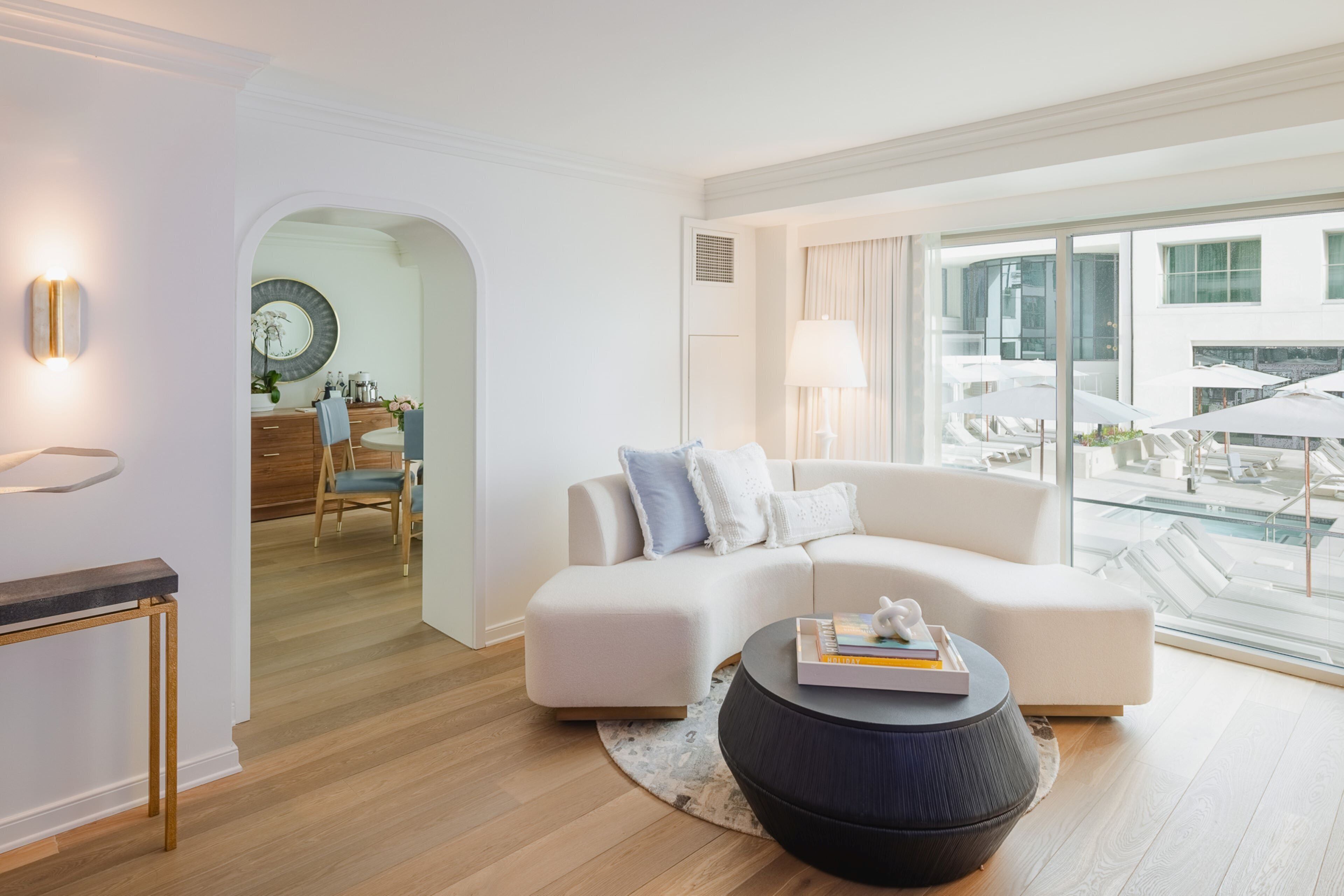 Una sala de estar moderna con un sofá blanco, una mesa redonda y vistas al área de la piscina a través de amplios ventanales.