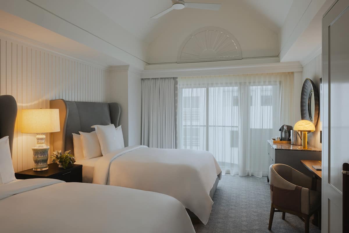 A hotel room with two beds, a desk, a chair, a ceiling fan, and a large window with curtains.