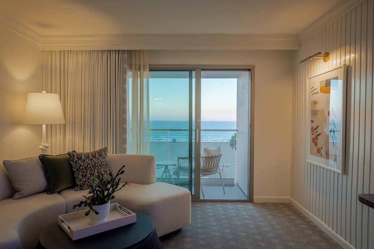 A hotel room with a sofa, a coffee table with a plant, a lamp, a sliding glass door to a balcony with a view of the sea, and a framed picture on the wall.