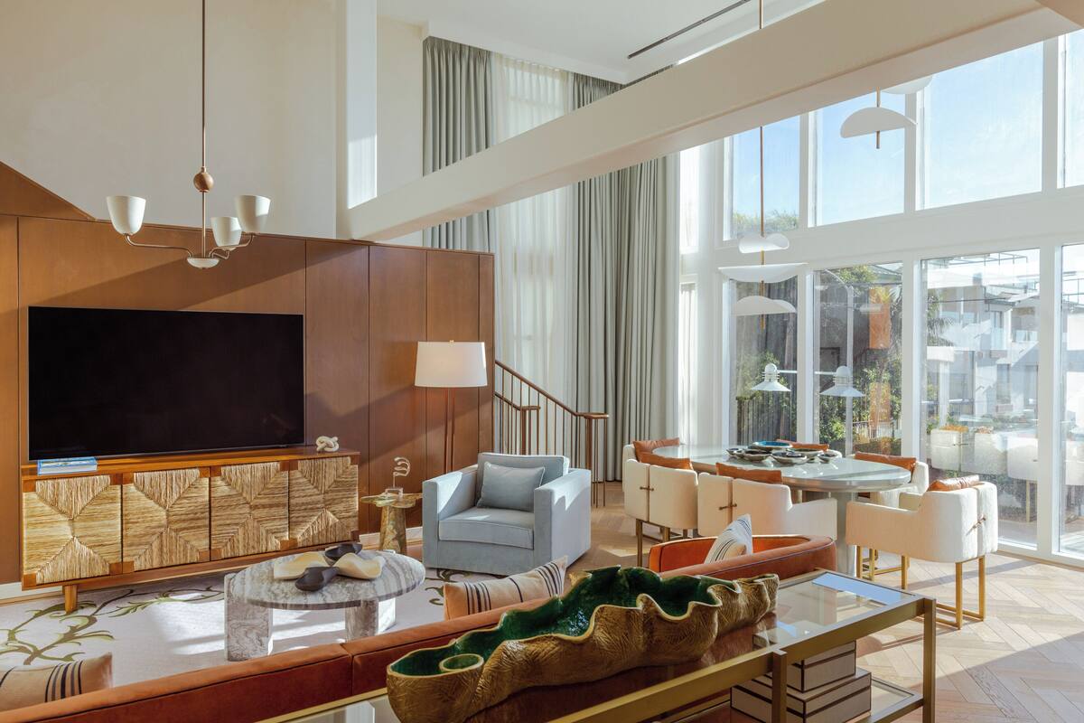 A spacious living area with a large flat-screen TV, comfortable seating, a dining table, and large windows offering a view of the city.