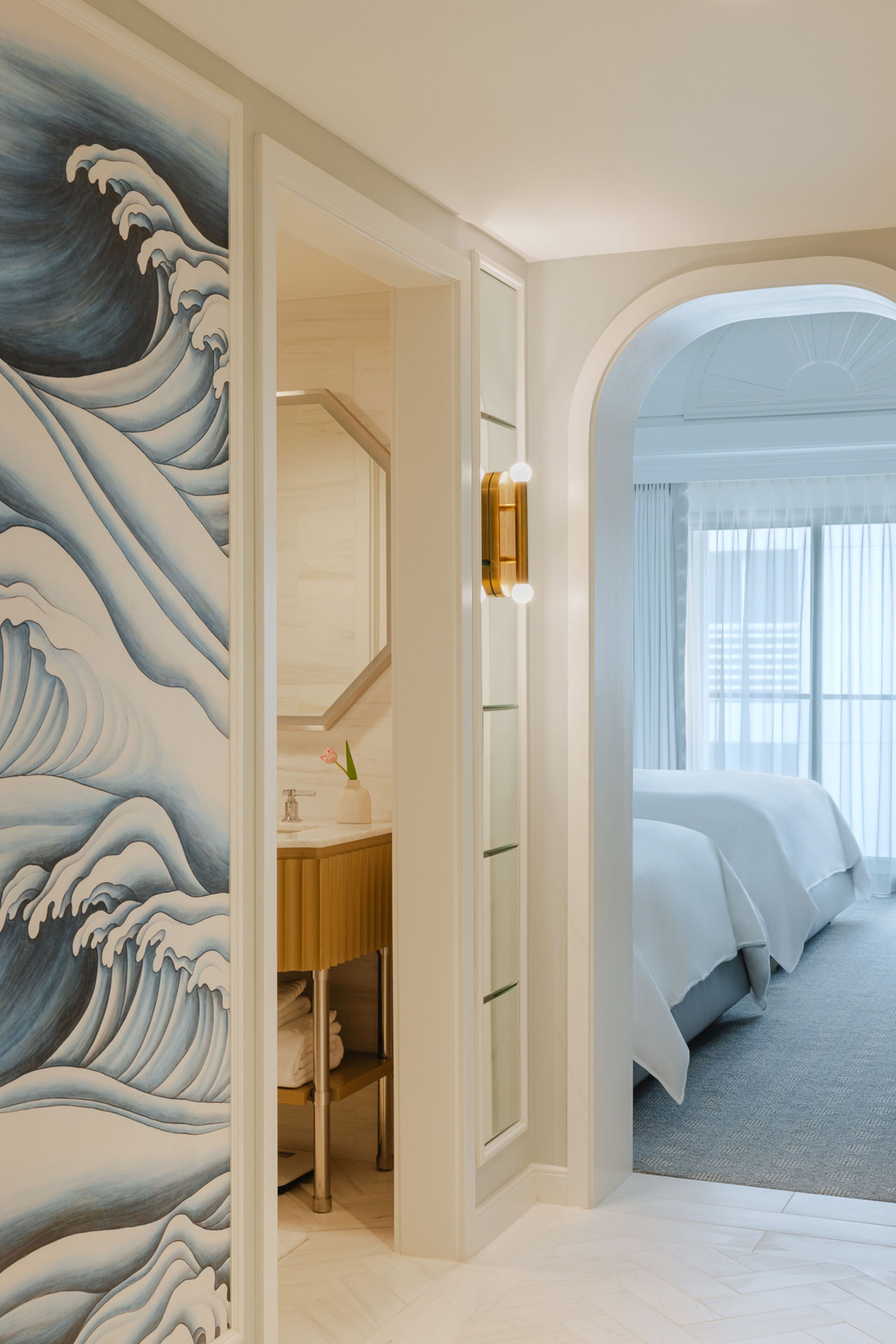 A hotel room with a wave painting on the wall, a bathroom with a sink and mirror, and a bed with white bedding.