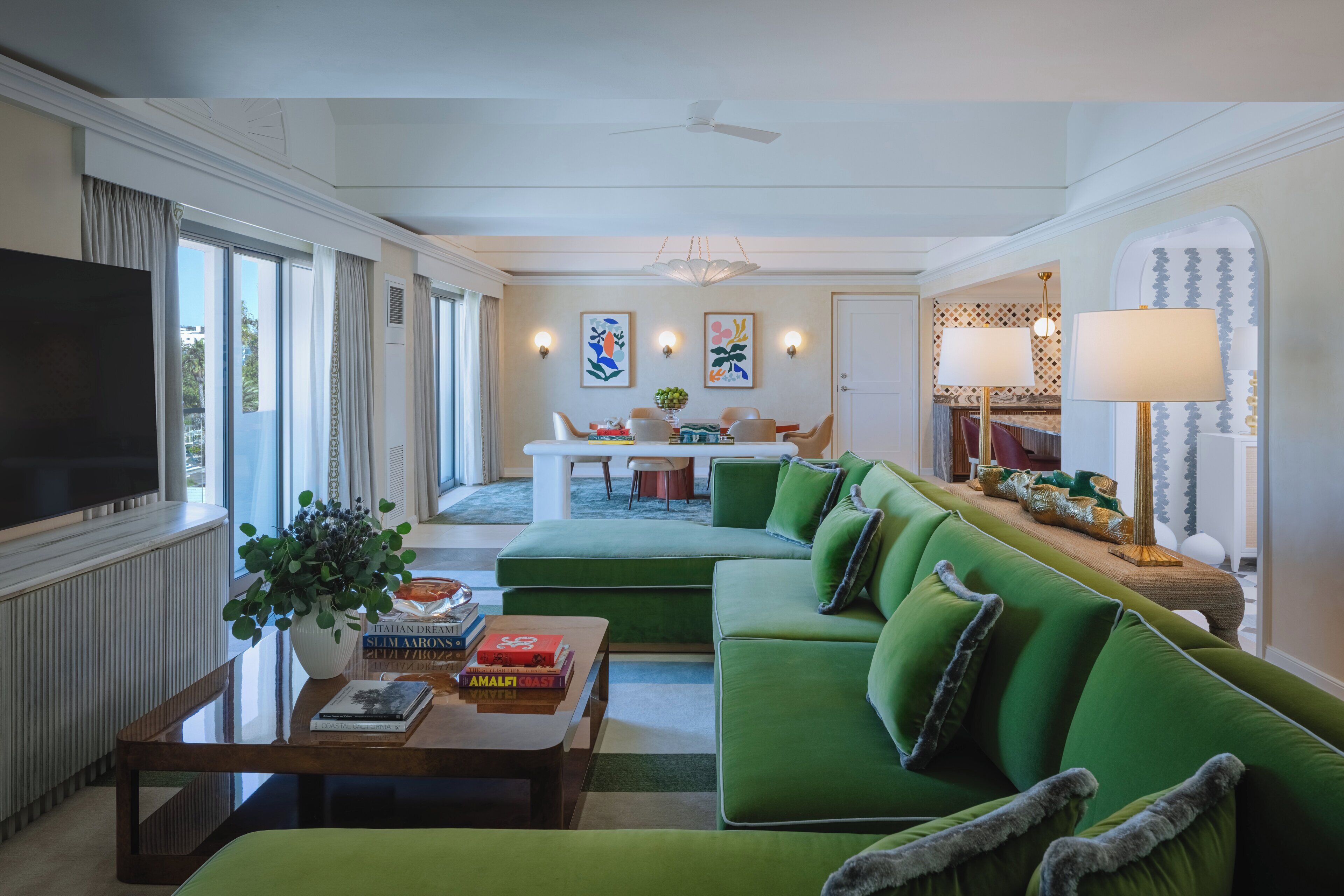 Un salon moderne avec un canapé vert, une table basse et un téléviseur. On aperçoit à l’arrière-plan un coin repas avec une table et des chaises blanches.