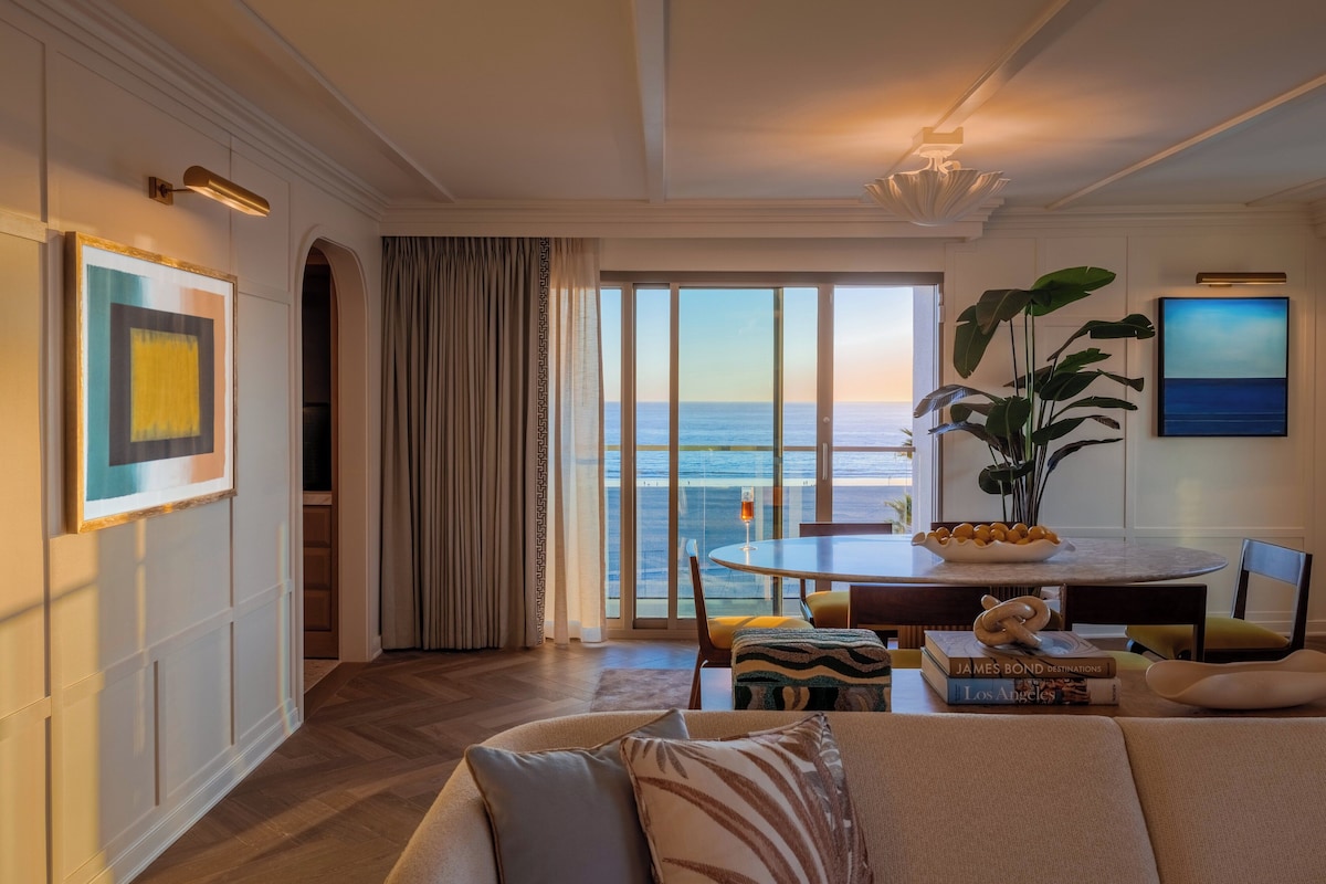 A modern living room with a large window overlooking the ocean, a dining area with a round table, and a cozy seating area with a sofa and cushions.
