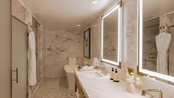 A modern bathroom with marble walls, a large mirror, dual sinks, and a walk-in shower.