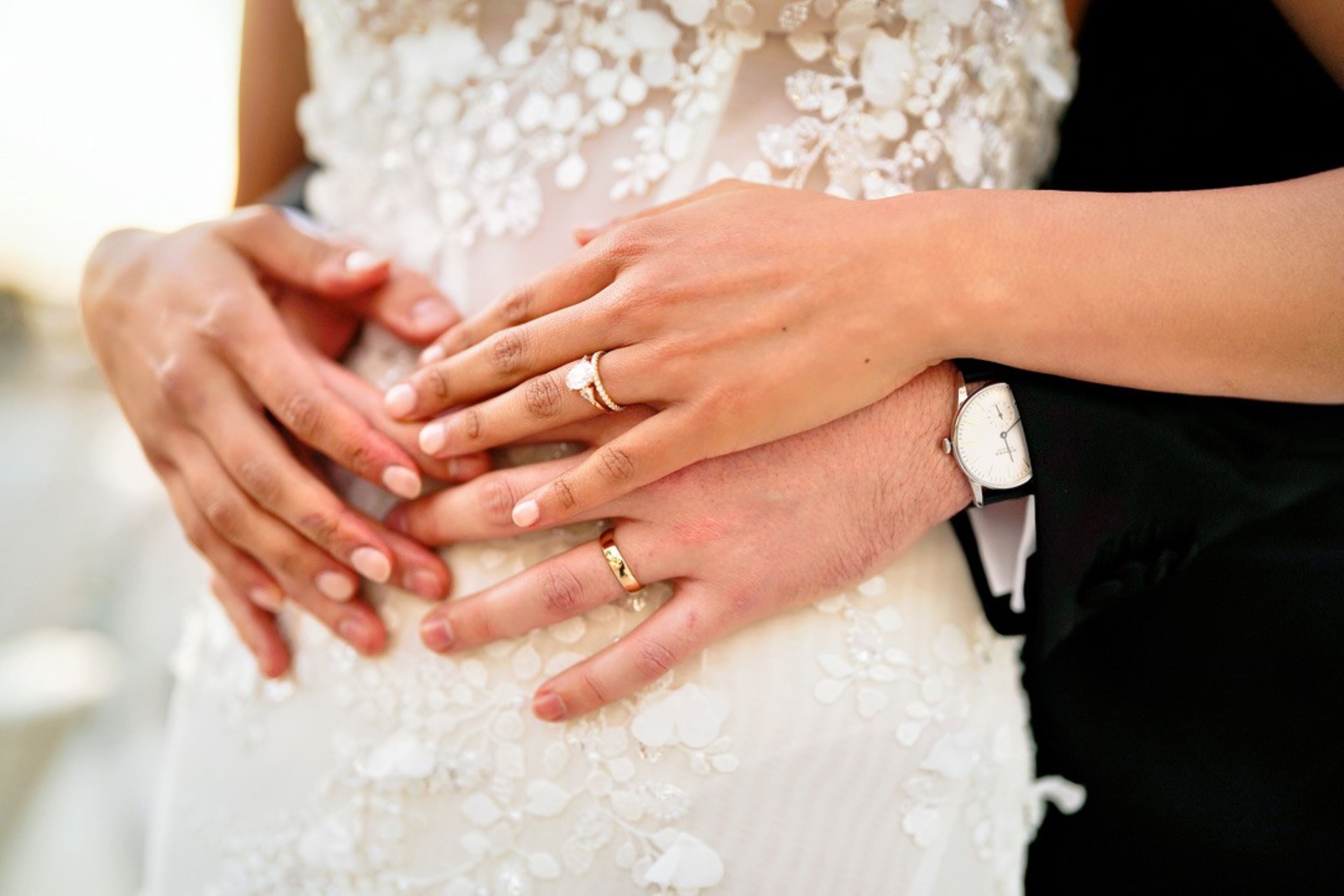 A close-up of hands, one wearing a wedding ring and the other a watch, on a white, floral-patterned garment.