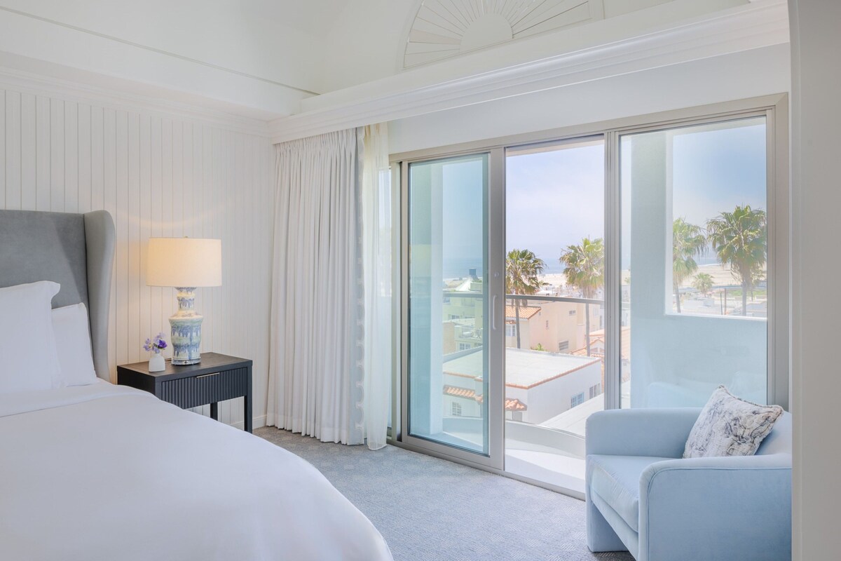 A hotel room with a large bed, a nightstand with a lamp, a blue armchair, and a sliding glass door leading to a balcony with a view of palm trees and buildings.