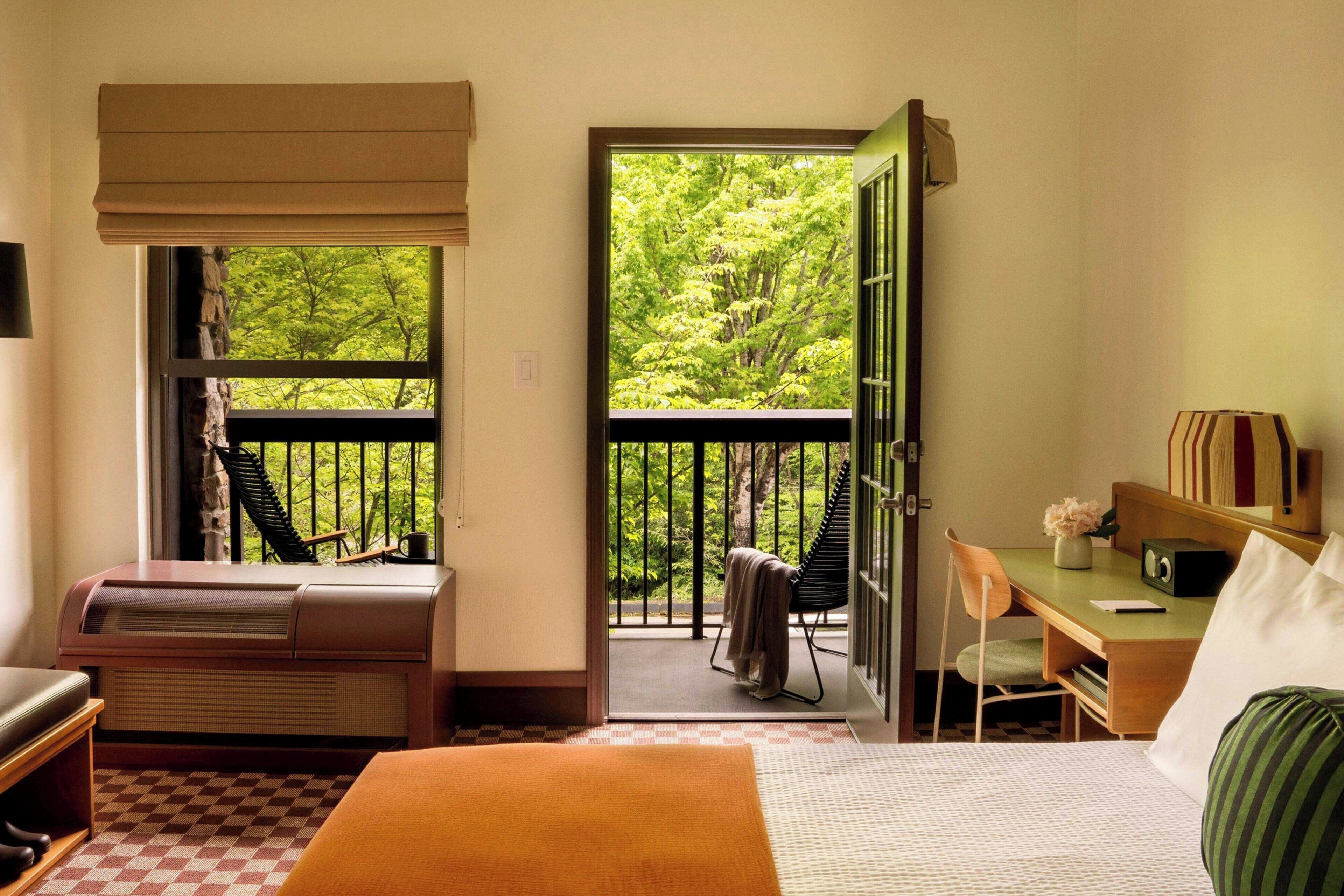 Une chambre d’hôtel comprenant un lit, un bureau, une chaise, un radiateur et un balcon avec vue sur les arbres.