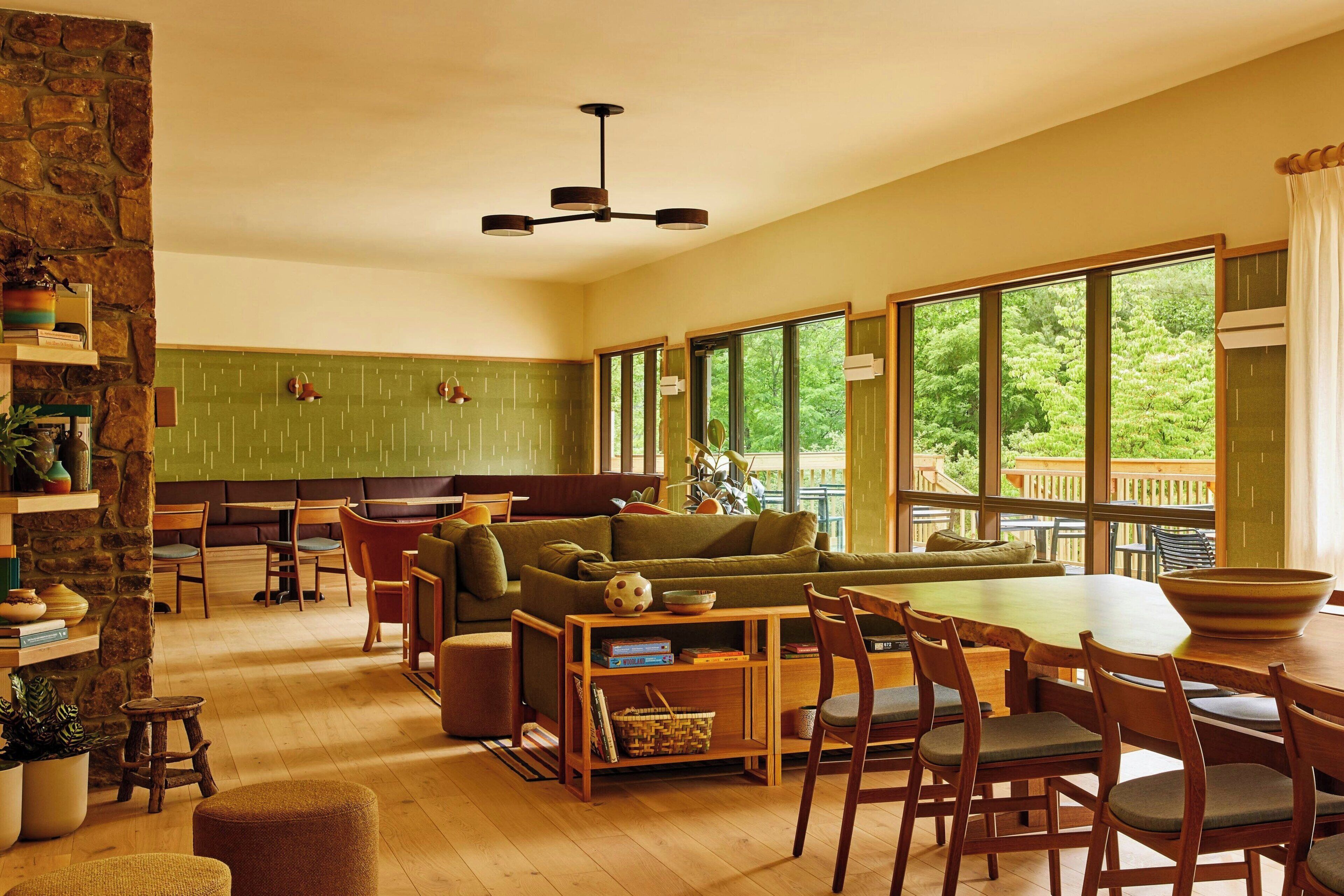 Ein gemütliches Wohnzimmer mit einem grünen Sofa, einem Holzespeisetisch und Stühlen. Eine Steinwand, eine hängende Leuchte und große Fenster mit Blick ins Grüne sind ebenfalls zu sehen.