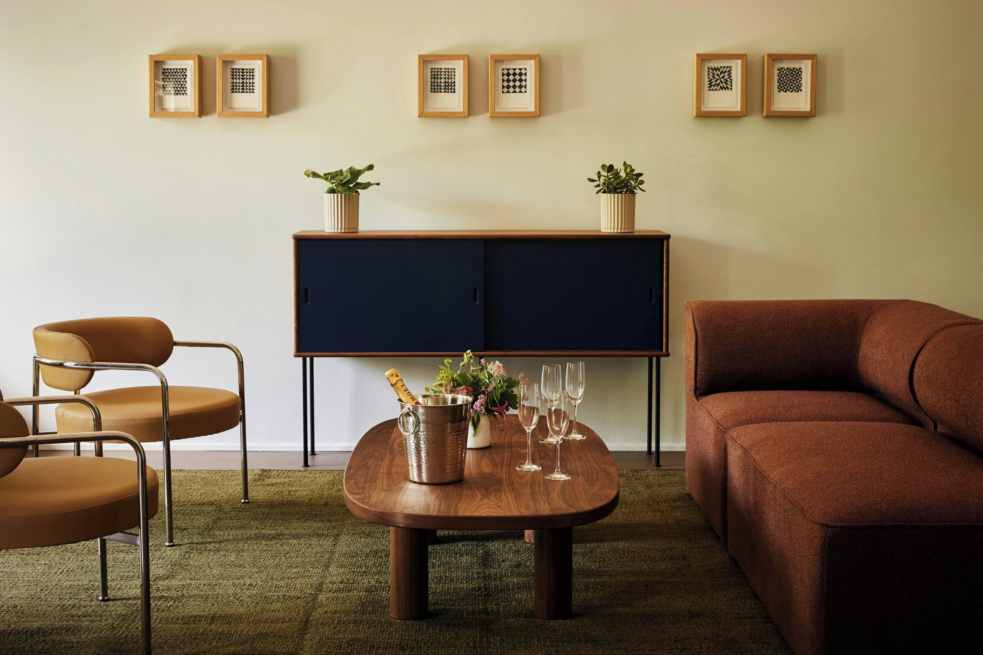 Una sala de estar con una mesa de centro de madera, un sofá marrón, un sillón marrón, un aparador azul oscuro y dos plantas en macetas.
