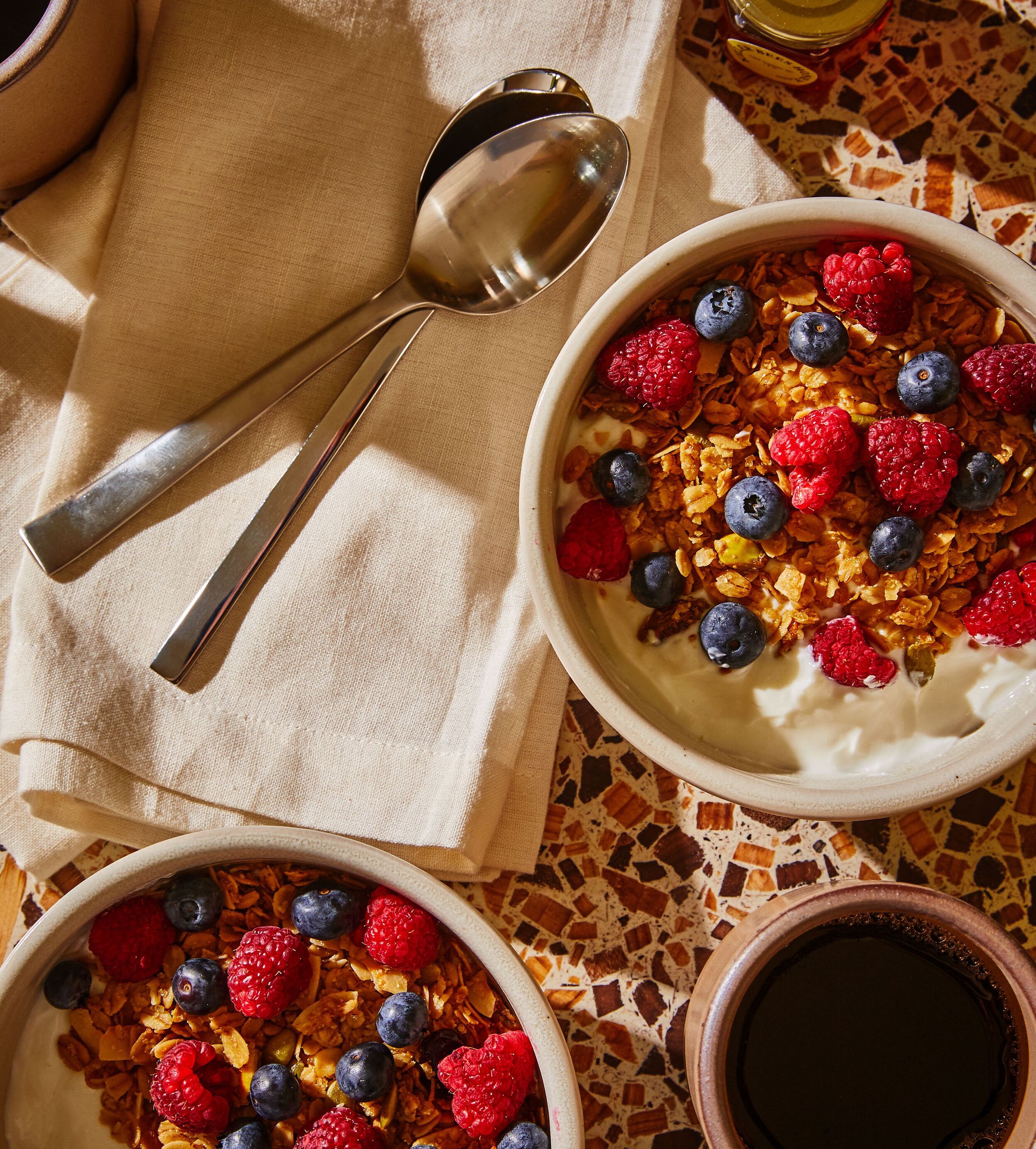 Deux bols de céréales avec des fruits rouges et du granola, une cuillère, une serviette et une tasse de café sur une table.
