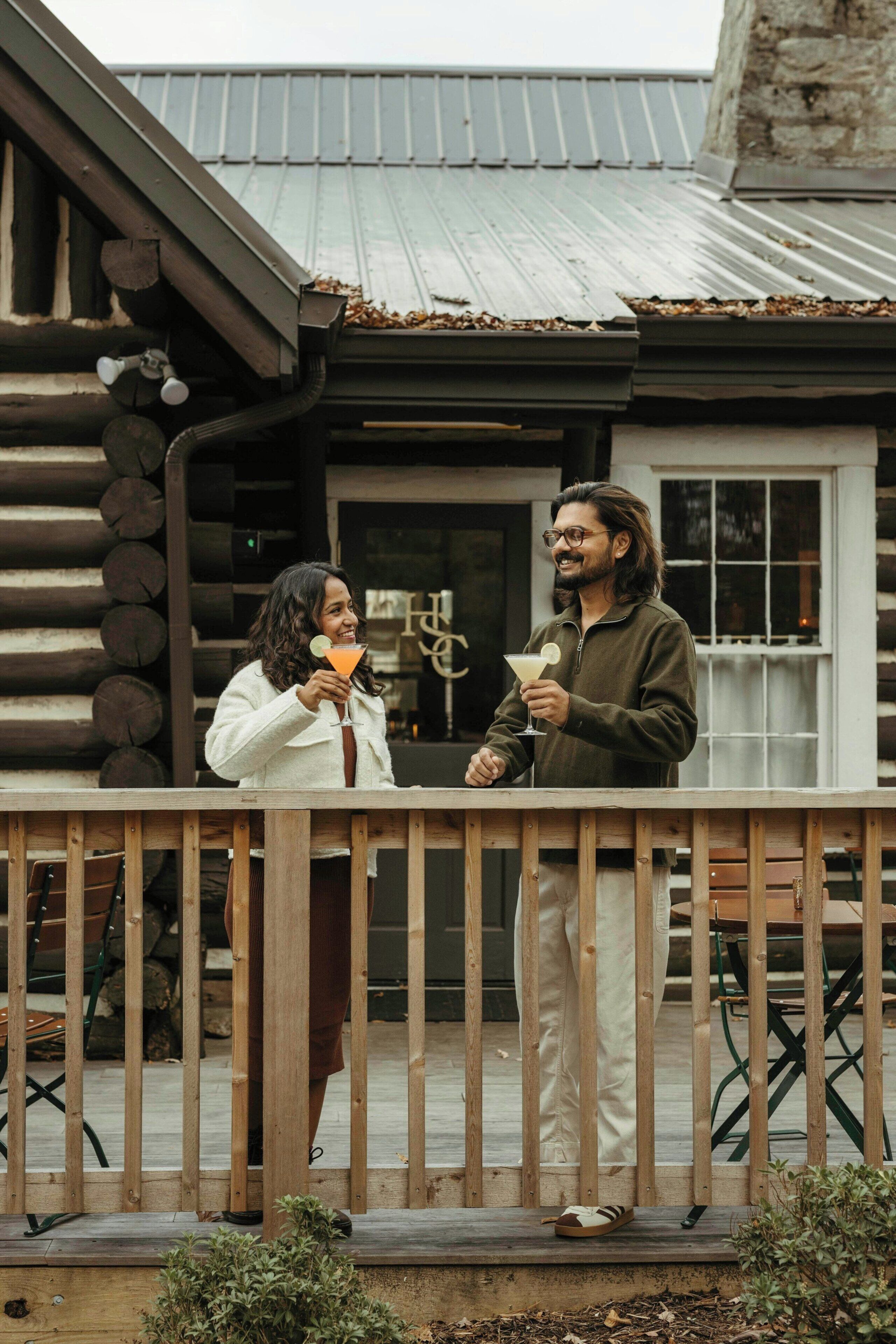 Two individuals standing on a wooden deck with a metal roof and stone walls in the background.