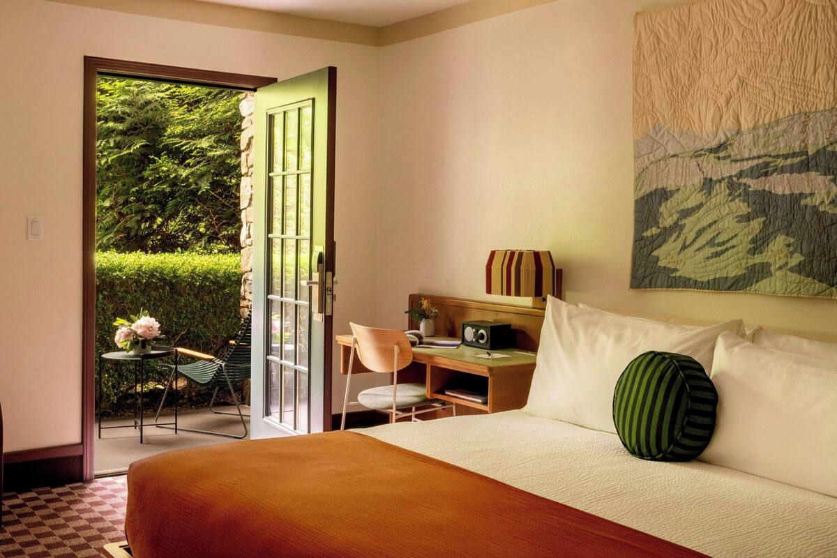 Une chambre avec un grand lit, un bureau et une chaise. Une porte donne sur un balcon d’où l’on peut admirer la verdure.