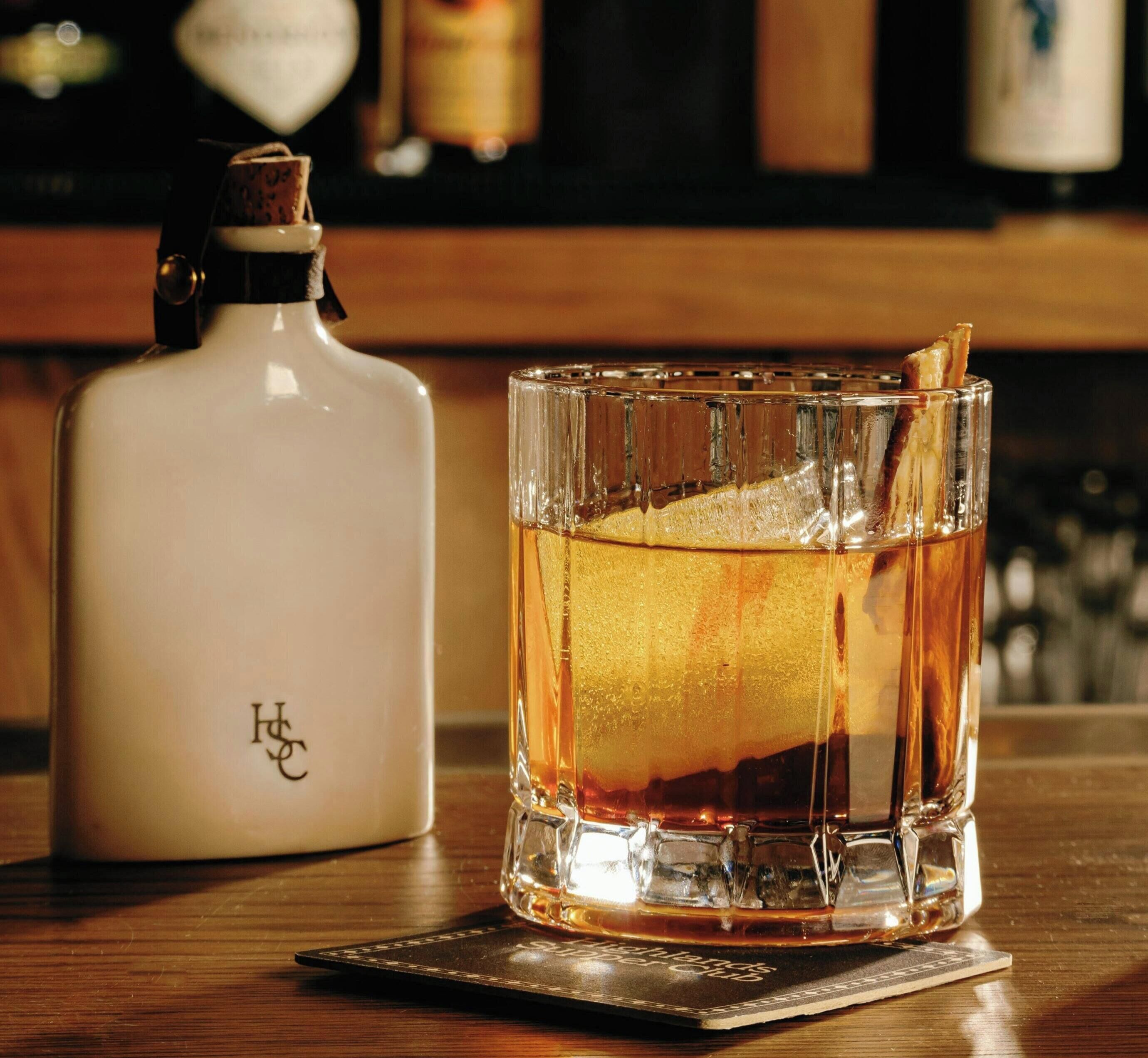 A glass of whiskey with a lemon peel garnish on a coaster, a bottle of whiskey, and a bar setting in the background.