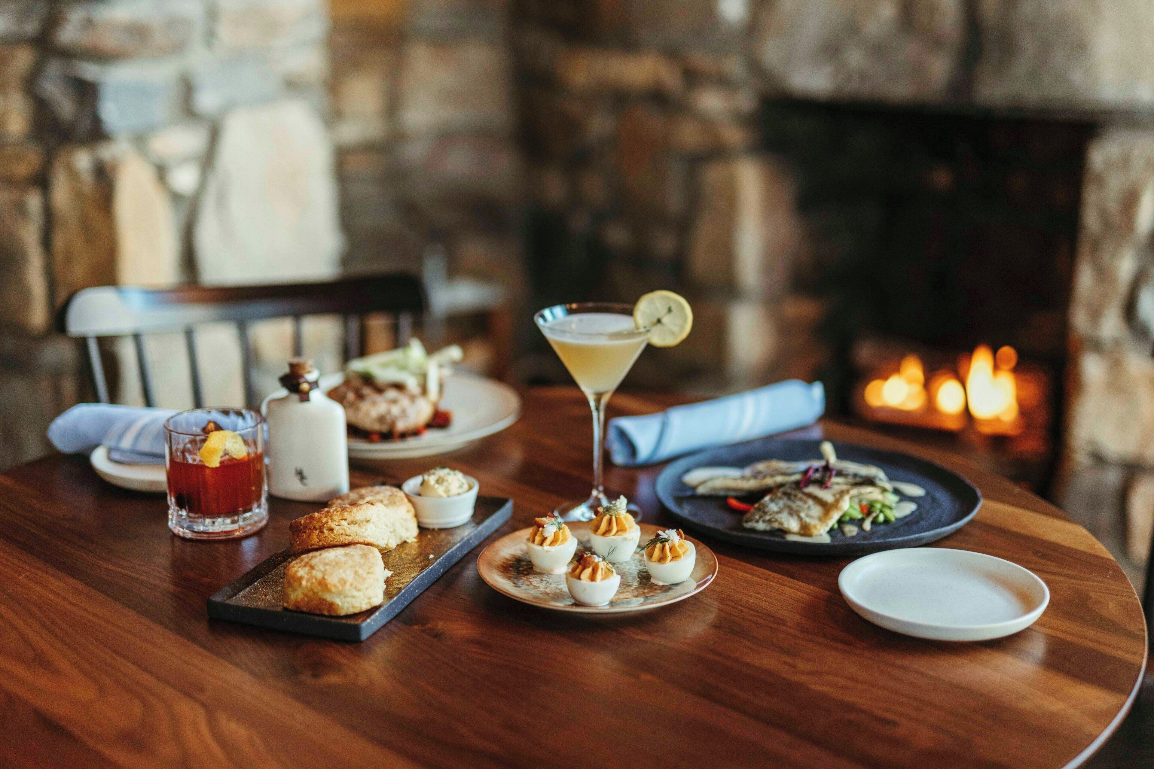 A wooden table with plates of food, a glass of red beverage, and a martini glass with a lemon garnish.